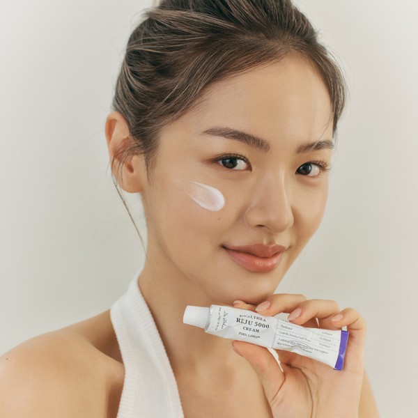 Modelo sostiene el tubo de Dr.Althea PDRN Reju 5000 Cream con crema aplicada en la mejilla.