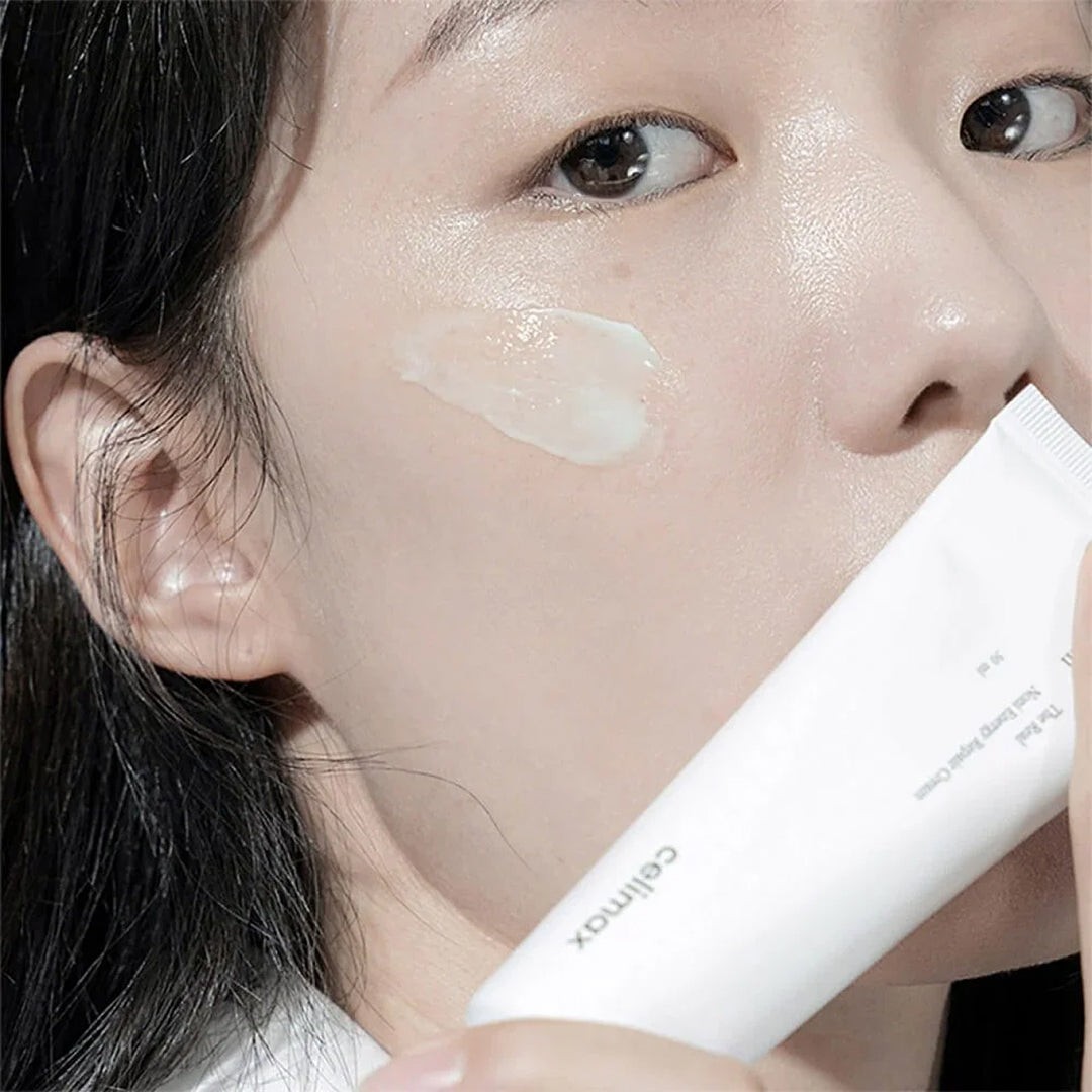 Modelo con crema aplicada en la mejilla sostiene el tubo de Celimax The Real Noni Energy Repair Cream.